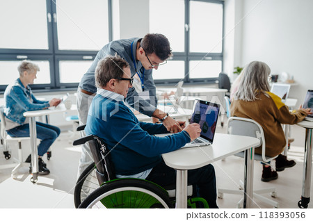 Teacher explaining senior man on wheelchair how to work with laptop. Elderly people attending computer and technology education class. Digital literacy. 118393056