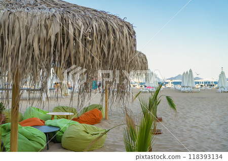 A beachside lounge area with colorful bean bags and thatched umbrella at sunset, serene seaside view A beachside lounge area with colorful bean bags and thatched umbrella at sunset, serene seaside view 118393134