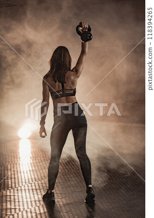 Empowered Fitness A determined woman confidently holding a kettlebell in a smokelit gym, radiating strength and focus, embodies the essence of hard work and dedication to fitness and wellbeing 118394265