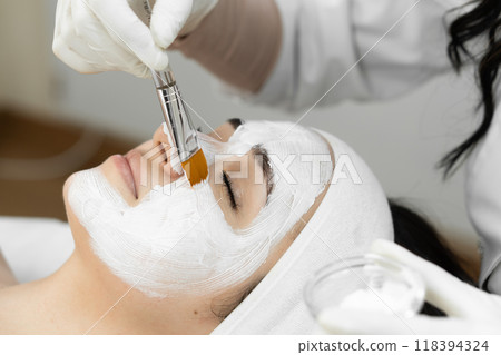Indulge in a luxurious facial treatment at a spa, where a beautician applies a white face mask for relaxation and skincare benefits. Rejuvenate and cleanse for ultimate wellbeing 118394324