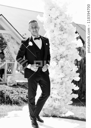 Elegant Groom in Stylish Black Tuxedo with Joyful Expression at Outdoor Wedding Ceremony with Floral Archway 118394700