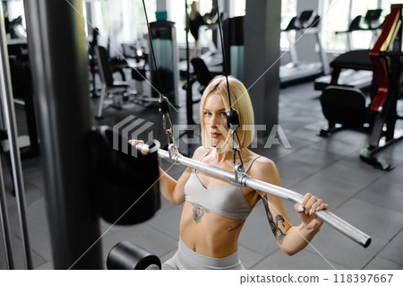 Sporty female woman in grey suit pulled on sports simulator in gym. Back muscles are very tense. Sporty female woman in grey suit pulled on sports simulator in gym. Back muscles are very tense. 118397667