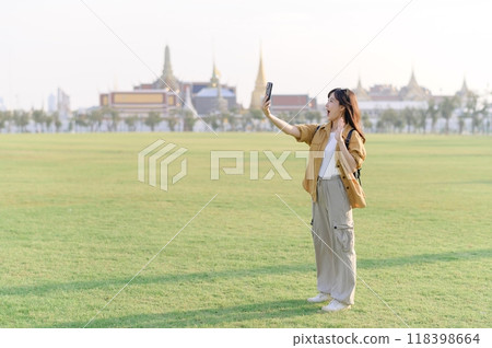 Traveler asian woman in her 30s take a livestream on smartphone while explores Wat Phra Kaew emerald Buddha. Share the wonders of Thai heritage through her journey. Traveler asian woman in her 30s take a livestream on smartphone while explores Wat Phra Kaew emerald Buddha. Share the wonders of Thai heritage through her journey. 118398664