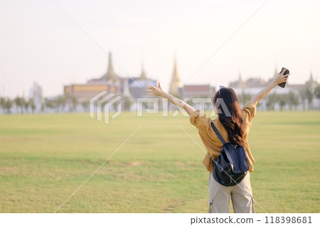 A Traveler Asian woman in 30s, bathed in golden glow of a Bangkok sunset, laughs with carefree joy, her arms outstretched as if to embrace freedom of the moment. Backpack strapped on. 118398681