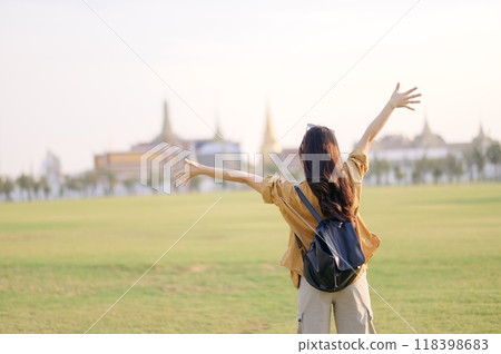 A Traveler Asian woman in 30s, bathed in golden glow of a Bangkok sunset, laughs with carefree joy, her arms outstretched as if to embrace freedom of the moment. Backpack strapped on. 118398683