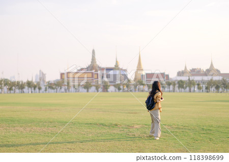 Traveler asian woman in her 30s, backpack slung over her shoulder, explores Wat Pra Kaew with childlike wonder. Sunlight dances on the golden rooftops, and a carefree smile 118398699