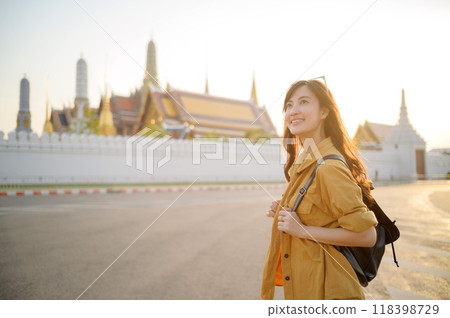 Traveler asian woman in her 30s, backpack slung over her shoulder, explores the intricate details of Wat Pra Kaew with childlike wonder. Sunlight dances on the golden rooftops. 118398729