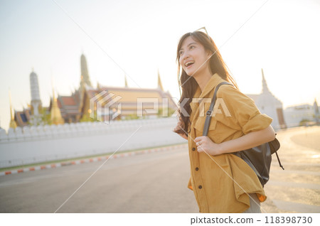 Traveler asian woman in her 30s, backpack slung over her shoulder, explores the intricate details of Wat Pra Kaew with childlike wonder. Sunlight dances on the golden rooftops. 118398730