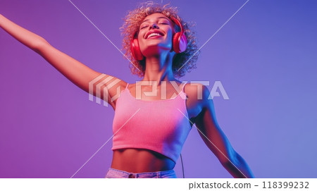 Energetic young woman with fair skin, curly blonde hair, and red headphones dancing against a purple gradient background in a pink tank top and high-waisted jeans. Happiness and lifestyle concept 118399232