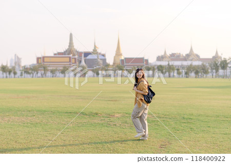 Traveler asian woman in her 30s, backpack slung over her shoulder, explores Wat Pra Kaew with childlike wonder. Sunlight dances on the golden rooftops, and a carefree smile Traveler asian woman in her 30s, backpack slung over her shoulder, explores Wat Pra Kaew with childlike wonder. Sunlight dances on the golden rooftops, and a carefree smile 118400922