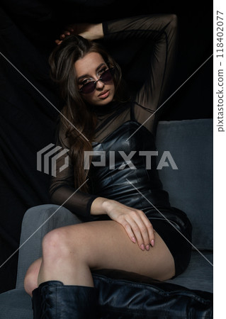 Beautiful fashionable woman in leather dress sitting on grey dark armchair 118402077