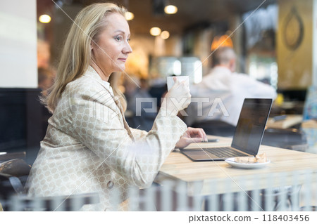 Business lunch - businesswoman sits at table in cafe by window with a laptop and drinks coffee. View through window Business lunch - businesswoman sits at table in cafe by window with a laptop and drinks coffee. View through window 118403456
