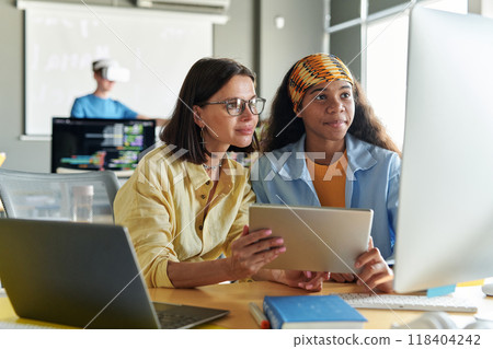 Young woman as female teacher using digital tablet and working with student at class in school or college while sitting at table with computer 118404242