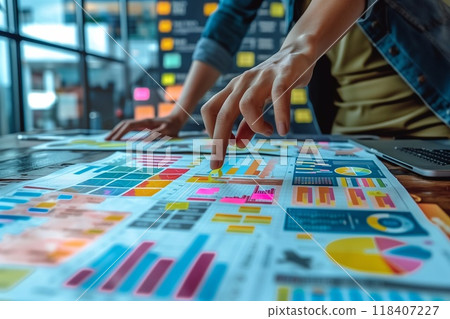 A hand points to a section of a chart on a large sheet of paper. There are other charts and graphs surrounding it, all with colorful bars and lines 118407227