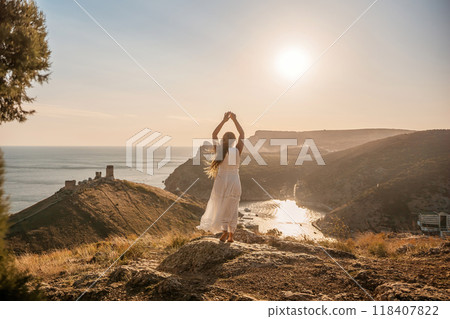 woman stands on a rocky hill overlooking a body of water. She is wearing a white dress and she is in a state of joy or celebration. Concept of freedom and happiness. 118407822