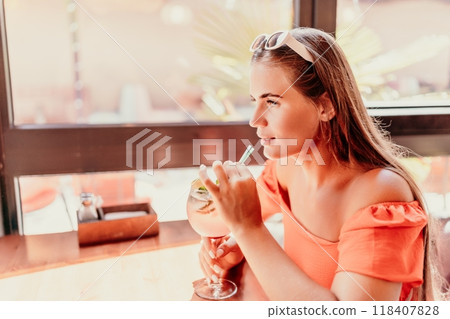 woman in a cafe seated, holding beverage. Dressed in an orange dress with her hair down. Bright interior, large windows allow natural light. Beverage consumption for refreshment. 118407828