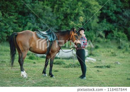 Happy blonde with horse in forest. Woman and a horse walking through the field during the day. Dressed in a plaid shirt and black leggings. 118407844