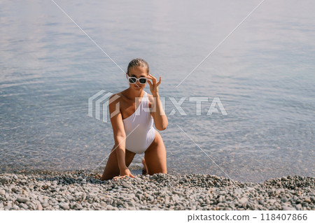 A woman in a white bikini is sitting on the beach, looking out at the water. She is wearing sunglasses and she is enjoying the view. A woman in a white bikini is sitting on the beach, looking out at the water. She is wearing sunglasses and she is enjoying the view. 118407866