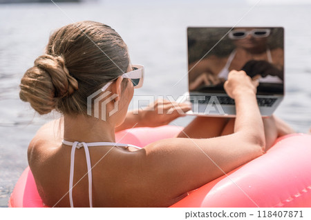 A woman is sitting on a pink inflatable raft in the ocean, reading a laptop. Concept of relaxation and leisure, as the woman enjoys her time by the water while working or browsing the internet. 118407871