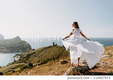 Happy woman in a white dress and hat stands on a rocky cliff above the sea, with the beautiful silhouette of hills in thick fog in the background. 118407873