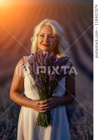 Blonde woman poses in lavender field at sunset. Happy woman in white dress holds lavender bouquet. Aromatherapy concept, lavender oil, photo session in lavender 118407874