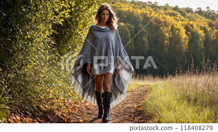 Fashionable woman in a stylish poncho leaves blowing boots stepping through foliage chic rural backdrop Fashionable woman in a stylish poncho leaves blowing boots stepping through foliage chic rural backdrop 118408279