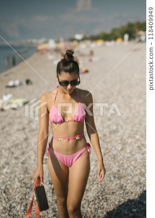 A young woman confidently walks along a beautiful pebble beach while wearing a pink bikini A young woman confidently walks along a beautiful pebble beach while wearing a pink bikini 118409949