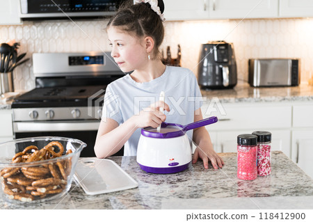 With focused attention, this budding culinary artist dips pretzels into a pot of melted chocolate, creating sweet delights in the warmth of a well-lit home kitchen. 118412900