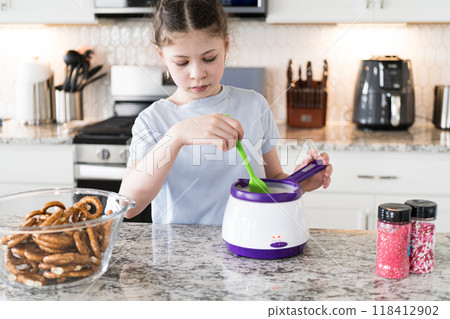 With focused attention, this budding culinary artist dips pretzels into a pot of melted chocolate, creating sweet delights in the warmth of a well-lit home kitchen. 118412902