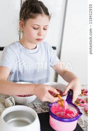 With focused attention, this budding culinary artist dips pretzels into a pot of melted chocolate, creating sweet delights in the warmth of a well-lit home kitchen. 118412910