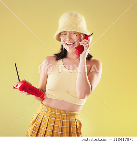 Landline, fashion and woman on a call in a studio with art eyes stickers on her face with 90s aesthetic. Communication, beauty and happy Asian female with a telephone isolated by a yellow background. Landline, fashion and woman on a call in a studio with art eyes stickers on her face with 90s aesthetic. Communication, beauty and happy Asian female with a telephone isolated by a yellow background. 118417875