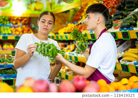 Polite young man seller of greengrocery shop helping woman client in choosing greenery 118419325
