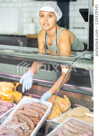 Positive young saleswoman demonstrating container with sausages in butcher shop Positive young saleswoman demonstrating container with sausages in butcher shop 118419343