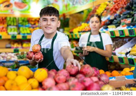 Young salesman proposing pomegranate, offering fruits in supermarket Young salesman proposing pomegranate, offering fruits in supermarket 118419401