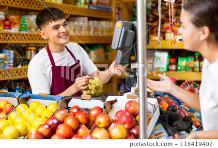 Friendly guy seller serving a girl customer weighs grapes on a scale Friendly guy seller serving a girl customer weighs grapes on a scale 118419402