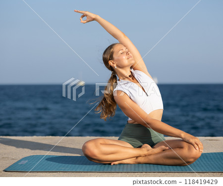 Young experienced woman fitness teacher performs yoga parshvottanasana sitting on beach at day 118419429