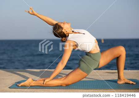 On windy day young slender woman does yoga near sea doing Crescent Moon pose On windy day young slender woman does yoga near sea doing Crescent Moon pose 118419446