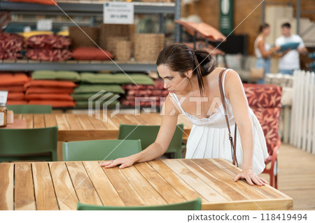 Pretty brunette girl in white dress chooses wooden table for country house in garden store Pretty brunette girl in white dress chooses wooden table for country house in garden store 118419494