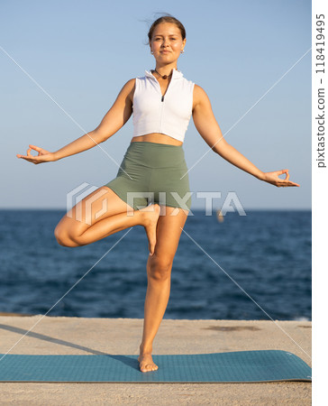 On windy day young slender woman does yoga near sea doing Vrikshasana 118419495