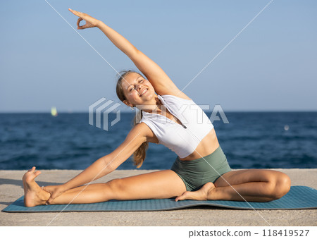 Young girl in sportwear doing stretching exercises for strong arms and abs and difficult yoga asana on the seashore on sunny day Young girl in sportwear doing stretching exercises for strong arms and abs and difficult yoga asana on the seashore on sunny day 118419527