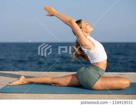 Sportive girl practicing pigeon pose of yoga on seashore Sportive girl practicing pigeon pose of yoga on seashore 118419531