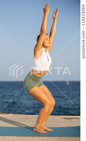 On windy day young slender woman does yoga near sea doing asana chair, utkatasana On windy day young slender woman does yoga near sea doing asana chair, utkatasana 118419569