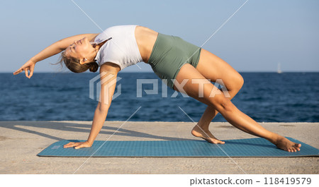 On windy day young slender woman does yoga near sea doing Chamatkarasana 118419579