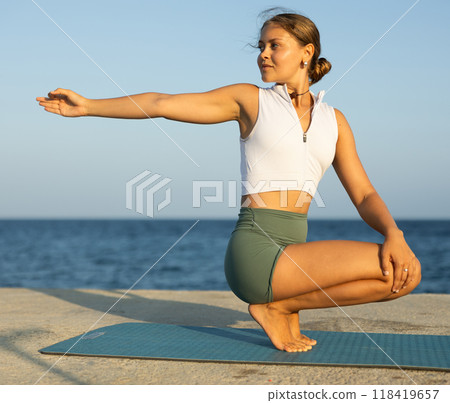 Young girl in sportwear doing stretching exercises for strong arms and abs and difficult yoga asana on the seashore on sunny day 118419657