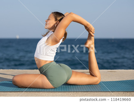 Flexible fit woman doing yoga on the seashore, performing seated back-bending asana Rajakapotasana, or King Pigeon Pose 118419660