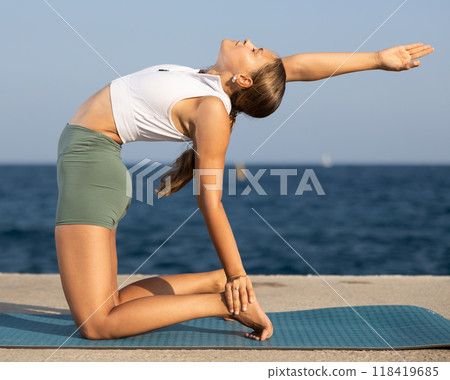 On windy day young slender woman does yoga near sea doing Ushtrasan 118419685