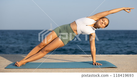 Positive girl practicing side plank pose of yoga on seashore Positive girl practicing side plank pose of yoga on seashore 118419693