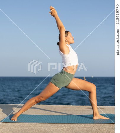 On windy day young slender woman does yoga near sea doing Virabhadrasana 118419770
