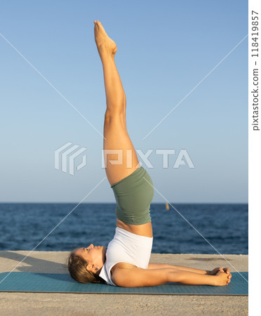 Sportive girl practicing shoulderstand pose of yoga on seashore Sportive girl practicing shoulderstand pose of yoga on seashore 118419857