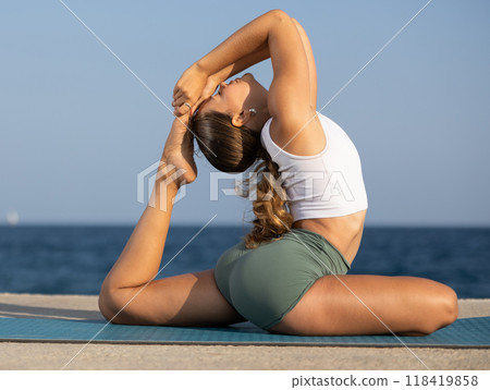 Flexible fit woman doing yoga on the seashore, performing seated back-bending asana Rajakapotasana, or King Pigeon Pose 118419858